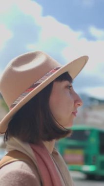 Dikey çekim. Güneşli bir havada şehrin arka planına karşı şapkalı neşeli beyaz bir kadının yakın plan portresi. Hafta sonu gezisi. Şapkalı şık kız kameraya gülümsüyor..
