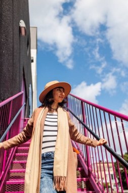 Şapkalı şık bir kız şehrin arka planına karşı korkulukların yanında poz veriyor. sokak tarzı ve moda.