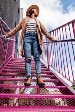 Şapkalı şık bir kız şehrin arka planına karşı korkulukların yanında poz veriyor. sokak tarzı ve moda.