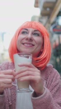 Dikey çekim. Turuncu saçlı güzel, mutlu bir kadın sokak kafesinde cappuccino içiyor. Mutlu kadın bir fincan kahve içip eğleniyor..