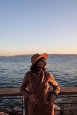 Paltolu ve şapkalı çekici genç bir kadının denize bakan portresi. Bir kız setin üzerinde duruyor ve denize bakıyor. sakinlik ve sükunet.