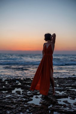 Gün batımında deniz kenarında poz veren kırmızı uzun elbiseli güzel, şehvetli bir kadın. Güzel gün batımı. Havada uçan bir kumaş şeridi..