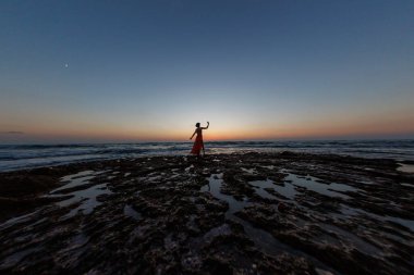 Gün batımında deniz kenarında poz veren kırmızı uzun elbiseli güzel, şehvetli bir kadın. Güzel gün batımı. Havada uçan bir kumaş şeridi..