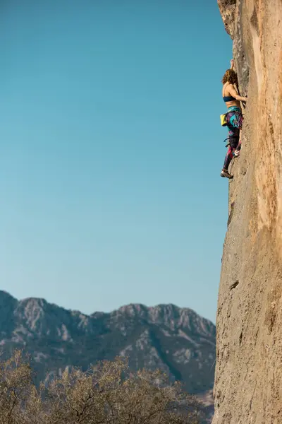 A woman climbs a rock against a blue sky, a strong girl trains strength and endurance, an extreme sport, rock climbing on natural terrain, a rock climber climbs with a rope