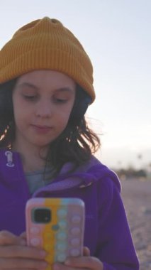 Dikey çekim. Kulaklık takan bir çocuk sahilde duruyor ve telefonundan dinlemek için bir şarkı seçiyor. Moda ve tarz. Sahilde ve arka planda dağlar