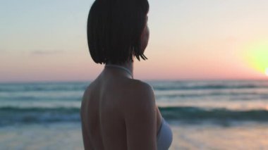 Gün batımının tadını çıkaran kadın. Beyaz mayolu ve bronzlaşmış tenli bir kız. Gün batımında sahilde poz vermek, dalgaların ve rahatlamanın tadını çıkarmak. Yaz havası.