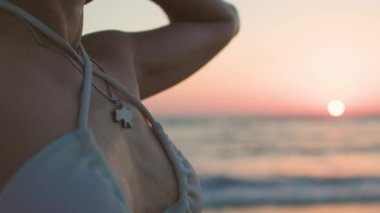 Gün batımının tadını çıkaran kadın. Beyaz mayolu ve bronzlaşmış tenli bir kız. Gün batımında sahilde poz vermek, dalgaların ve rahatlamanın tadını çıkarmak. Yaz havası.