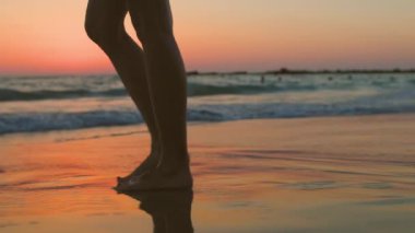 Gün batımında bir kız kumsalda yalınayak yürüyor, kumda ayak izleri ve suda yansımalar bırakıyor. Sahilde kadın bacaklarına yakın çekim. Sakinlik ve sükunet. Yaz havası.