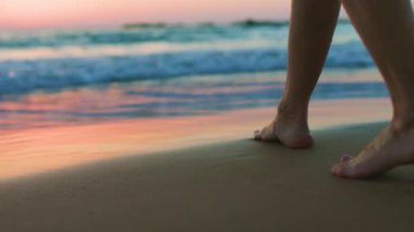 Gün batımında bir kız kumsalda yalınayak yürüyor, kumda ayak izleri ve suda yansımalar bırakıyor. Sahilde kadın bacaklarına yakın çekim. Sakinlik ve sükunet. Yaz havası.