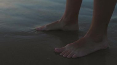 Gün batımında bir kız kumsalda yalınayak yürüyor, kumda ayak izleri ve suda yansımalar bırakıyor. Sahilde kadın bacaklarına yakın çekim. Sakinlik ve sükunet. Yaz havası.