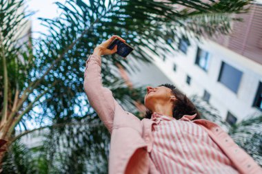 Neşeli aktif kadın güzel bir şehir parkının fotoğrafını çekiyor. Şehir manzarasının fotoğrafını çekmek için modern akıllı telefonu kullanan mutlu bir kız. Çekici turist kız dışarıda fotoğraf çekmeyi seviyor.