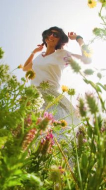 Dikey çekim. Yaz havası. Mavi gökyüzü ve çiçeklerle dolu bir arka planda poz veren genç ve neşeli bir kız. Çiçeklerin arasında mutlu bir kız ve bahar doğası dışarıda dinleniyor. bahar modası.