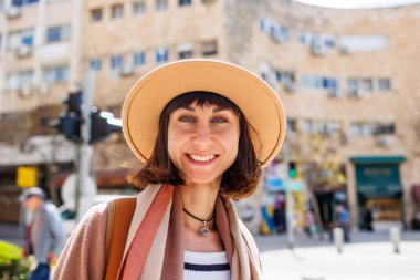 Güneşli bir havada şehrin arka planına karşı şapkalı neşeli beyaz bir kadının yakın plan portresi. Hafta sonu gezisi. Kameraya gülümseyen şapkalı şık bir kızın fotoğrafı..