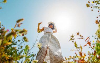 Yaz havası. Mavi gökyüzü ve çiçeklerle dolu bir arka planda poz veren genç ve neşeli bir kız. Güzel bir kız çiçekler arasında dans eder. Çiçeklerin arasında mutlu bir kız, açık havada. bahar modası.