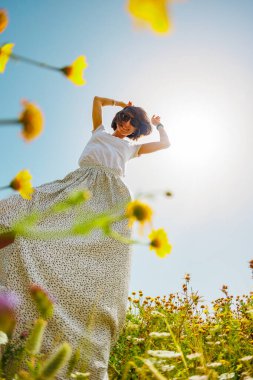 Yaz havası. Mavi gökyüzü ve çiçeklerle dolu bir arka planda poz veren genç ve neşeli bir kız. Çiçeklerin arasında dans eden kız. bahar modası.