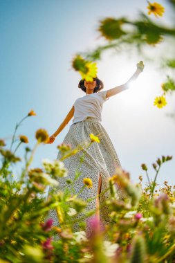 Yaz havası. Mavi gökyüzü ve çiçeklerle dolu bir arka planda poz veren genç ve neşeli bir kız. Güzel bir kız çiçekler arasında dans eder. Çiçeklerin arasında mutlu bir kız, açık havada. bahar modası.