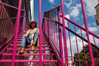 Şehirde merdivenlerde otururken şapkalı şık bir kız poz veriyor. Moda tarzı.