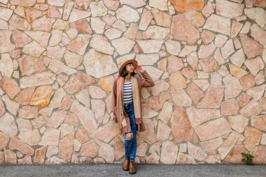 Şık giyinmiş ve şapkalı güzel bir kız. Sokakta poz veren kaygısız seksi kadın. Açık havada esmer bir model. Neşeli ve mutlu. Duvarın yanında..