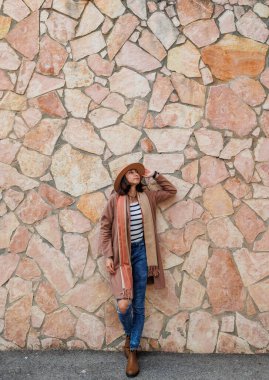 Güzel, gülümseyen bir modelin portresi. Bir şehir duvarının arkasında şapka takıyor ve şık kıyafetler giyiyor. Bir kadın sokakta bir duvarın yanında poz veriyor. Komik ve pozitif kadın dışarıda eğleniyor.. 