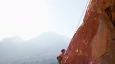 Dağcı çocuk. Yaz kampında spor. Bir çocuk yaz kampında kaya tırmanışı yapıyor. Güneşli bir günde doğada spor. Sevimli genç, kayaya bağlanmış bir şekilde tırmanıyor. Aktif tatil. Yaz tatili.