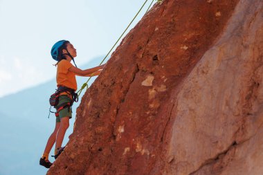 Yaz kampında spor. Bir çocuk yaz kampında kaya tırmanışı yapıyor. Dağcı çocuk. Güneşli bir günde doğada spor. Sevimli genç, kayaya bağlanmış bir şekilde tırmanıyor. Aktif tatil. Yaz tatili.