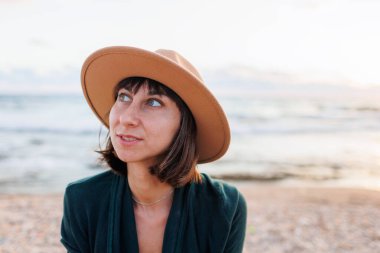 Güzel, doğal, güzel bir kadın gülümseyip gülüyor, kumsalda kameraya poz veriyor. Şapkalı gülümseyen genç bir kızın portresi. Kumsalda oturan mutlu bir gezgin..