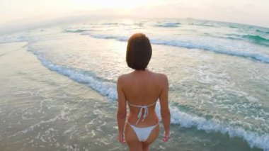 Sahilde yaz günü. Arkadan bak. Beyaz mayo giymiş genç bir kadın kumlu bir sahil boyunca okyanusa doğru yürüyor, günbatımının arkaplanına karşı. Kız tatilinin tadını çıkarıyor.