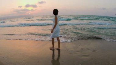 Beyaz elbiseli mutlu kadın plajda dinlenmeyi, gün batımında kumlu sahilde yürümeyi seviyor. Kumsaldaki mutlu kız. Kız sahilde yalınayak yürüyor..