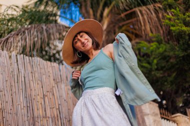 Genç bir esmer şehirde dururken mutlu bir şekilde gülümsüyor. Gömlekli, etekli ve şapkalı şık bir kız avlunun yanında duruyor. Yaz Portresi