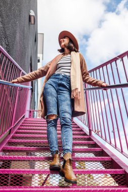 Şapkalı şık bir kız şehrin arka planına karşı korkulukların yanında poz veriyor. sokak tarzı ve moda.