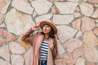 Güzel, gülümseyen bir modelin portresi. Bir şehir duvarının arkasında şapka takıyor ve şık kıyafetler giyiyor. Bir kadın sokakta bir duvarın yanında poz veriyor. Komik ve pozitif kadın dışarıda eğleniyor.. 