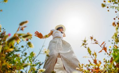 Yaz havası. Mavi gökyüzü ve çiçeklerle dolu bir arka planda poz veren genç ve neşeli bir kız. Güzel bir kız çiçekler arasında dans eder. Çiçeklerin arasında dans eden kız. bahar modası.