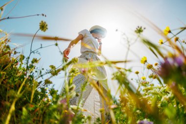 Yaz havası. Mavi gökyüzü ve çiçeklerle dolu bir arka planda poz veren genç ve neşeli bir kız. Çiçeklerin arasında dans eden kız. bahar modası.