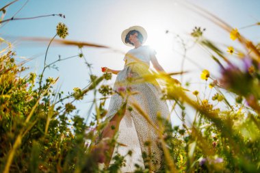 Yaz havası. Mavi gökyüzü ve çiçeklerle dolu bir arka planda poz veren genç ve neşeli bir kız. Çiçeklerin arasında mutlu bir kız ve bahar doğası dışarıda dinleniyor. bahar modası.