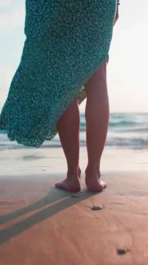 Dikey çekim. Yürüyüşün tadını çıkar. Dişi bacaklar yakın plan. Arkadan bak. Ayaklarını suya sok. Elbiseli bir kadın sahilde durup gün batımını seyrediyor. Sahilde yalınayak duran bir kız.