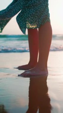 Dikey çekim. Dişi bacaklar yakın plan. Yan görünüş. Dalgalı elbiseli bir kadın sahilde duruyor. Sahilde çıplak ayakla duran bir kız. Sahilde gün batımı. Yürüyüşün tadını çıkar.