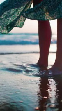 Dikey çekim. Dişi bacaklar yakın plan. Yan görünüş. Dalgalı elbiseli bir kadın kıyıda duruyor, kumda ayak izleri ve suda yansımalar bırakıyor. Sahilde gün batımı. Yürüyüşün tadını çıkar.