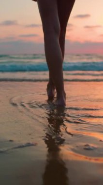 Dikey çekim. Sahilde yürüyüşün tadını çıkarıyorum. Bacaklarını kapat. Mayo giymiş ince bir kadın gün batımında sahilde yürüyor. Gün batımında bir kız sahilde yalınayak yürüyor..