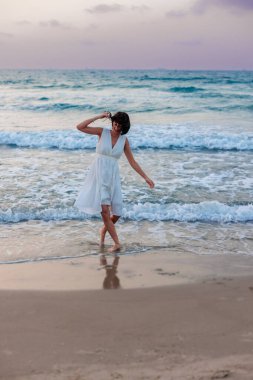 Beyaz yazlık elbiseli mutlu genç kız gün batımında sahilde yürüyor. Kumsalda dans eden kız..