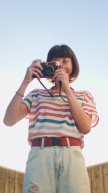 Dikey çekim. Genç bir kız kamerayı yüzüne tutar ve ateş eder. Elinde analog film kamerası olan şık ve çekici bir kadın. seyahat ve tatil.