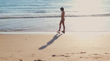 Beyaz mayo giymiş genç ve narin bir kadın kumlu sahil boyunca yürüyor..