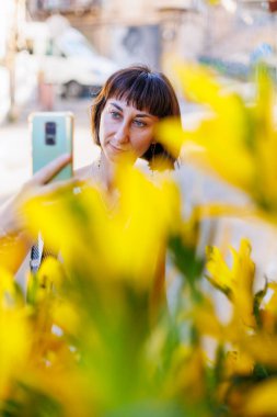 Akıllı telefon kullanan genç bir kadın dışarıda dekoratif bir çiçeğin fotoğrafını çekiyor. Güneşli bir günde güzel bir bitkinin fotoğrafını çeken bir kadının elleri mobil bir cihaz tutuyor. Şehirde dolaş..