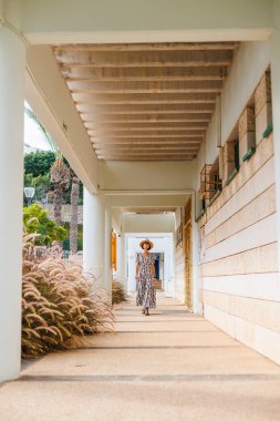 Şapka takan güzel bir bayanın tam boy fotoğrafı. Sokakta yürümek, güneşli hava, hafta sonu. Elbiseli ve şapkalı genç bir kız sokakta yürüyor..