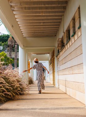 Şapka takan güzel bir bayanın tam boy fotoğrafı. Sokakta yürümek, güneşli hava, hafta sonu. Elbiseli ve şapkalı genç bir kız sokakta yürüyor..