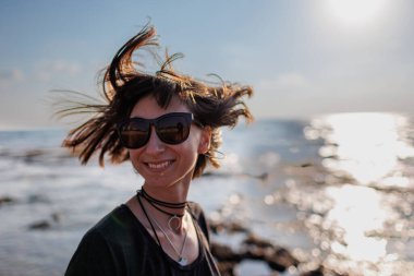 Saçları rüzgarda uçuşan genç bir kız. Sahilde poz veren şık ve neşeli bir kızın portresi. Neşeli hafta sonu.
