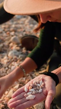 Dikey çekim. Genç bir kız sahilde oturur ve farklı kabuklar toplar. Deniz kabuğu koleksiyonu. Kızın elleri, yakın plan kabuklu. dinlenme ve meditasyon.