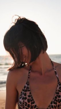 Dikey çekim. Kumsalda güzel bir esmer. Genç mutlu kız gülümsüyor. Saç yüzünü kapatır. Rüzgar saçlarınla oynuyor. Yüz kılları. Yaz tropik havası.