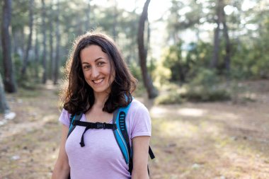 Sırt çantalı bir kadın. Vahşi doğadaki doğa faaliyetlerinin arka planında orman ve dağların yaşam tarzı macera konsepti. Genç kız kameraya gülümsüyor..