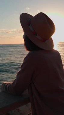 Dikey çekim. Paltolu ve şapkalı çekici genç bayan denize bakıyor. Gün batımında yürü. Kız denize bakmaktadır. sakinlik ve sükunet.