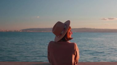 Paltolu ve şapkalı çekici genç bayan denize bakıyor. Gün batımında yürü. Kız denize bakmaktadır. sakinlik ve sükunet.